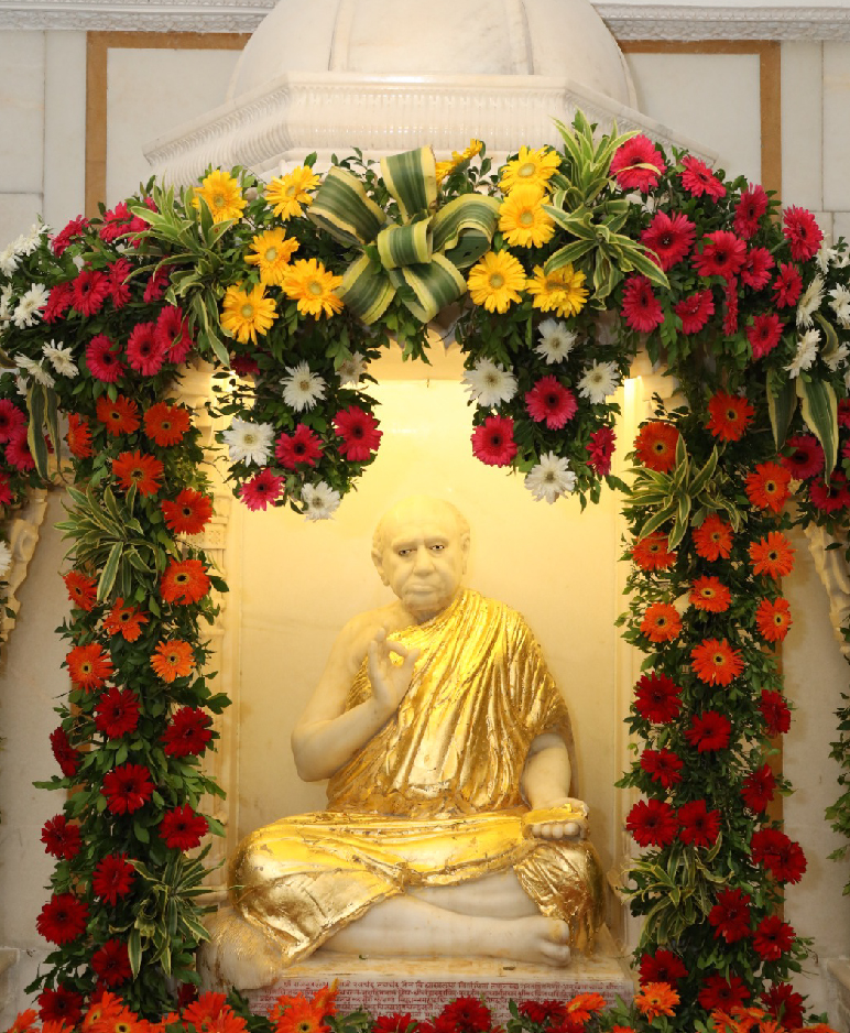 A statue of Bapji Maharaj in a Floral background with flowers around the statue.