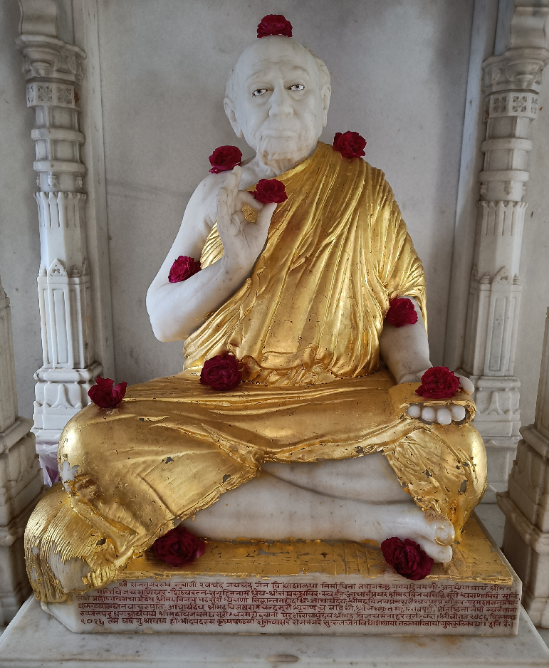 White marble statue of a Bapji Maharaj holding a pink flower, with other flowers nearby.